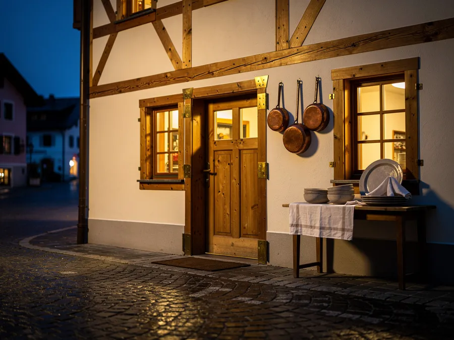 Fachwerk-Gasthaus in der blauen Stunde mit Holztür, warm beleuchteten Fenstern und Kupferpfannen an der Fassade; davor ein Tischchen mit Tellern und eine leere, nasse Pflasterstraße.