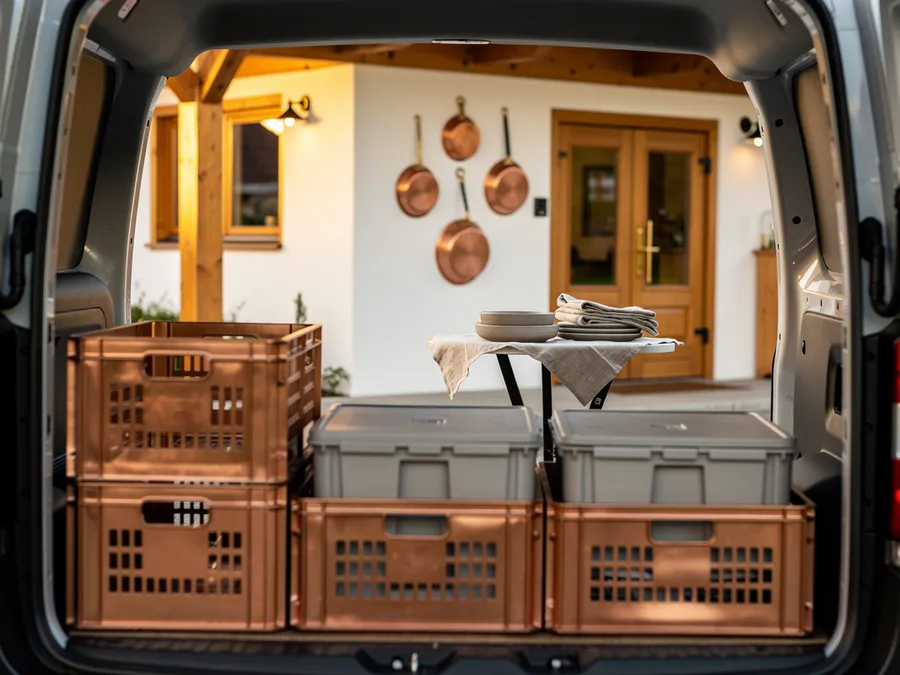 Blick aus dem Laderaum eines Transporters auf kupferfarbene Kisten und graue Thermoboxen; dahinter der Eingang eines Gasthauses mit Kupferpfannen an der Wand und ein Tischchen mit Geschirr.
