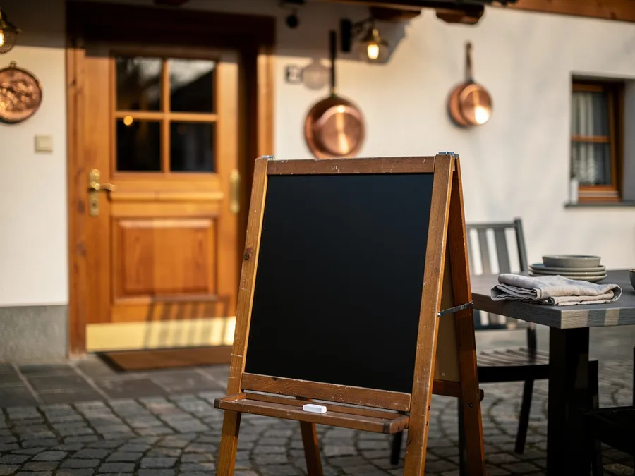 Leere schwarze Kreidetafel mit einem Stück Kreide auf einem Holzaufsteller vor der Eingangstür eines Gasthauses; daneben ein Tisch im Freien mit Geschirr und Serviette.